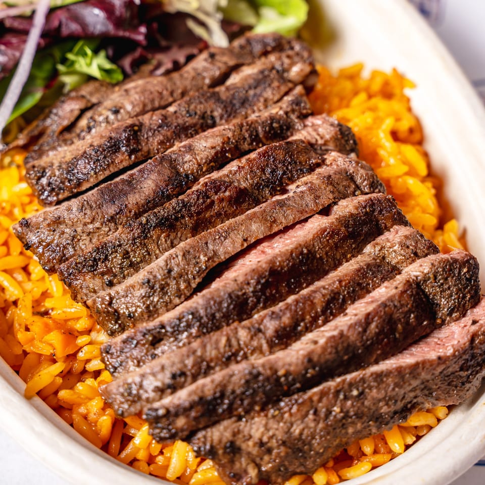 Grilled steak slices over yellow rice with a side salad, served in a white bowl.