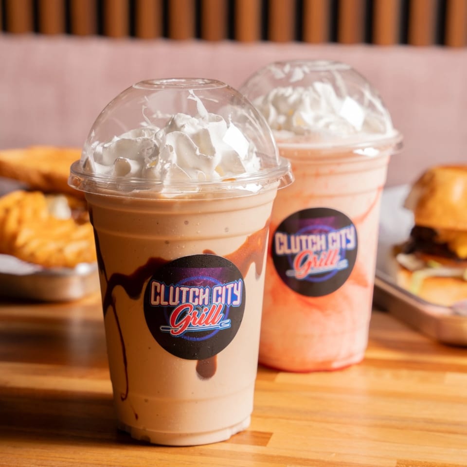 Two Clutch City Grill milkshakes, one chocolate and one strawberry, with whipped cream and food in the background.