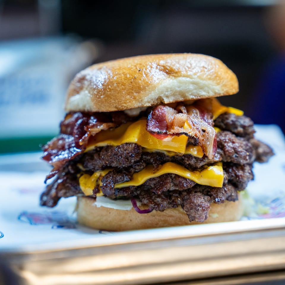 A towering triple-patty burger from Clutch City Grill, loaded with melted cheese and crispy bacon.