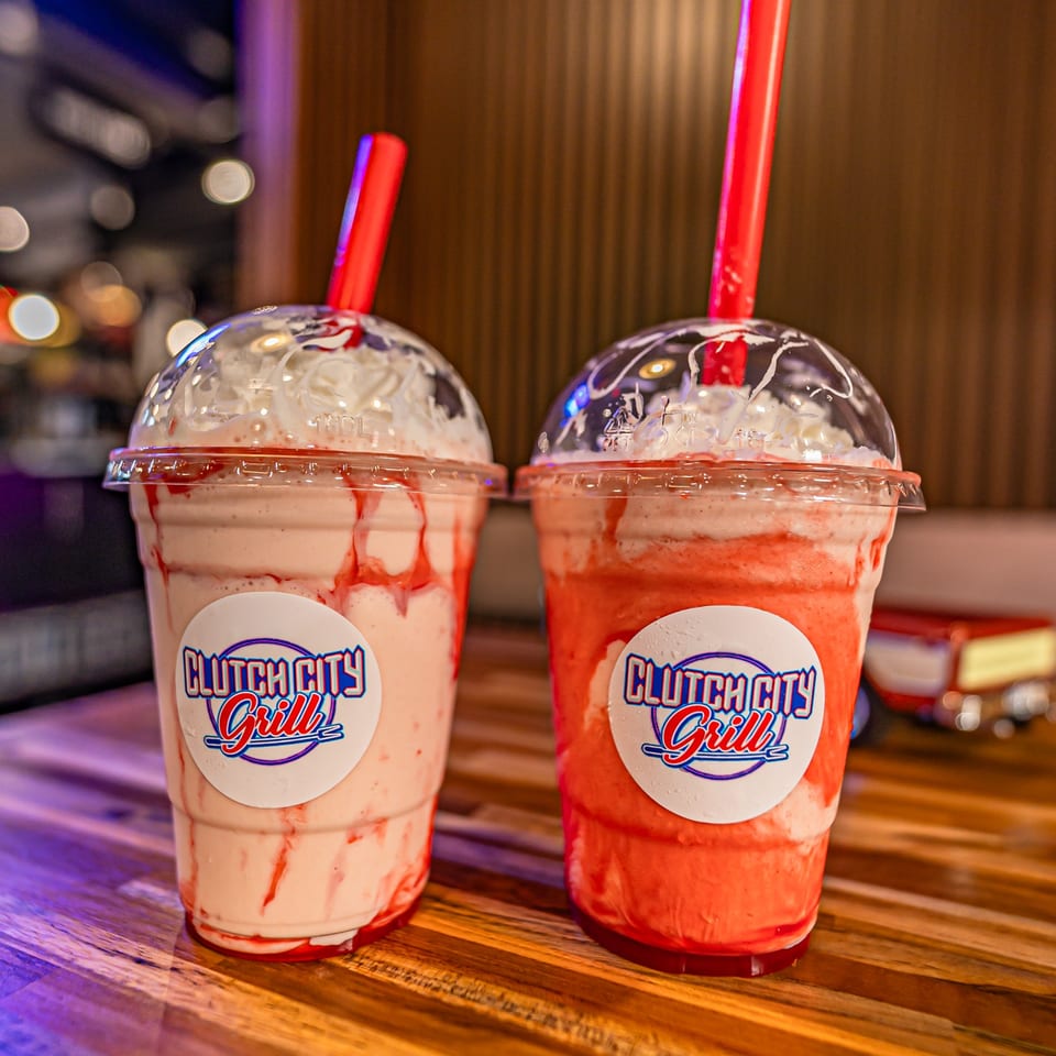 Two strawberry milkshakes with whipped cream and red straws, featuring Clutch City Grill logos.