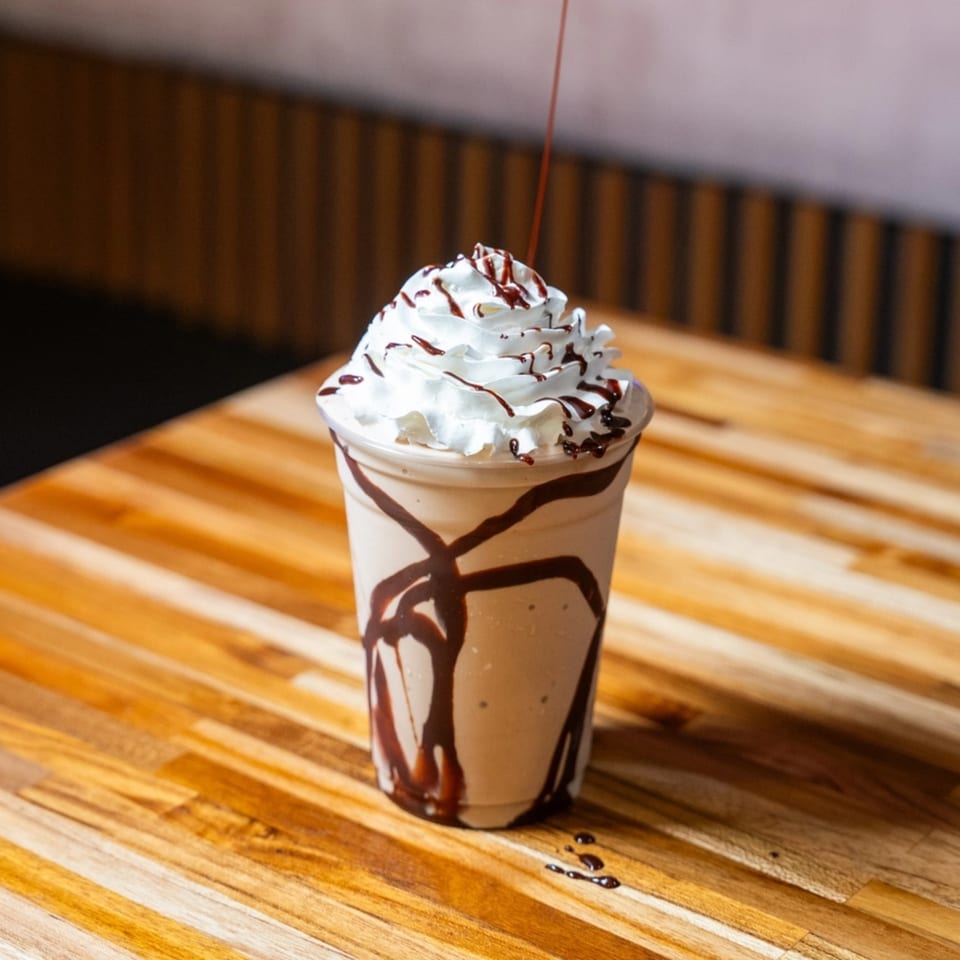 A chocolate milkshake with whipped cream and chocolate drizzle sits on a wooden table at Clutch City Grill.