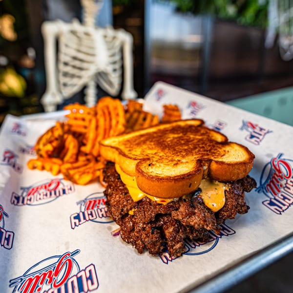 Clutch City Grill's patty melt with waffle fries on branded paper, with a skeleton in the background.