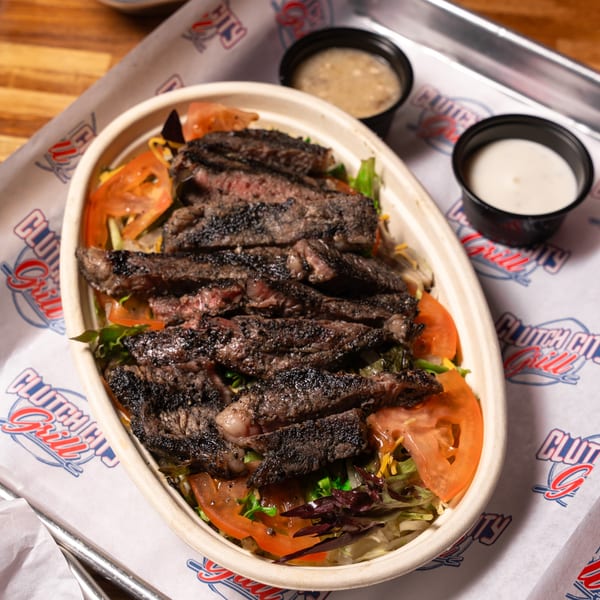 Grilled steak salad with tomatoes and lettuce, served with two dressing cups on Clutch City Grill branded paper.