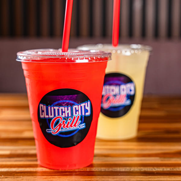Two Clutch City Grill drinks, one red and one yellow, with red straws on a wooden table.