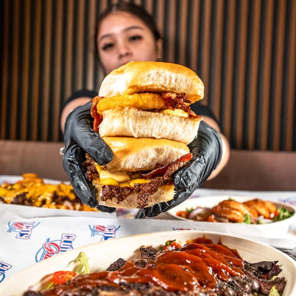 A person in black gloves holds a towering burger with bacon and hash browns at Clutch City Grill.