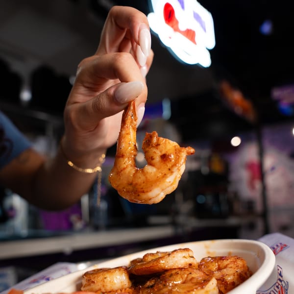 A hand holds up a seasoned shrimp from a bowl of other shrimp at Clutch City Grill.