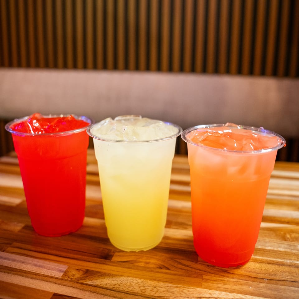 Three colorful iced drinks, red, yellow, and orange, on a wooden table at Clutch City Grill.