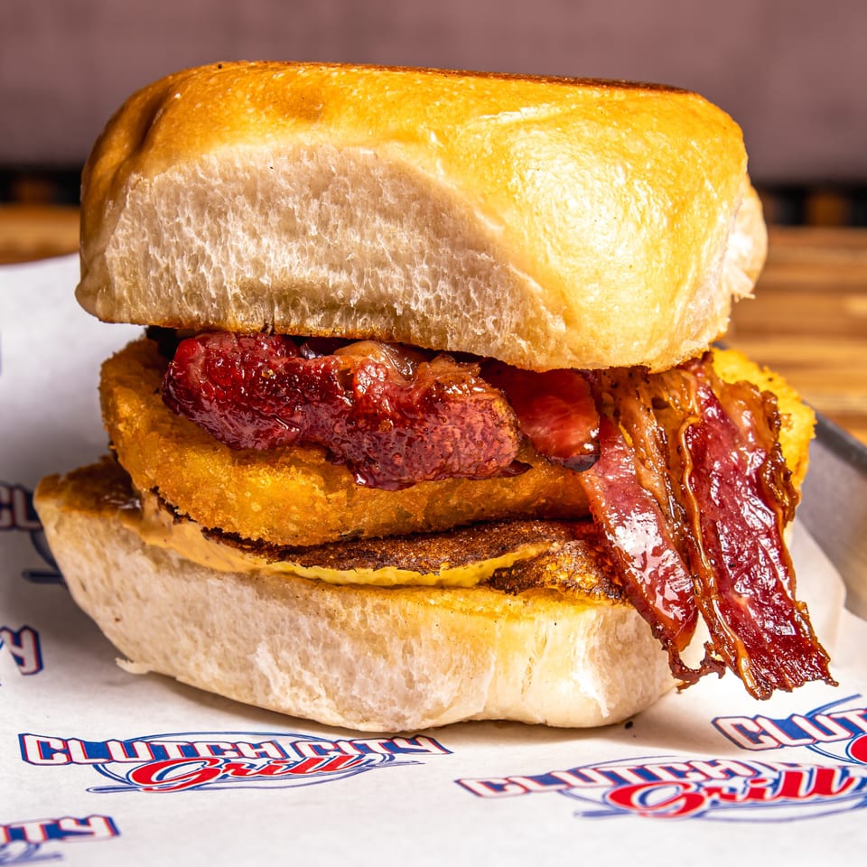 Close-up of a Clutch City Grill breakfast sandwich with bacon, hash brown, and egg on a bun.