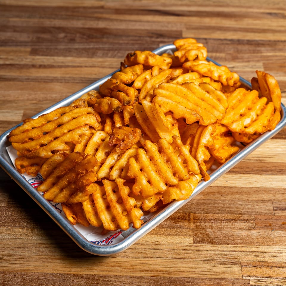 A metal tray overflowing with golden-brown waffle fries, seasoned with spices, on a wooden table.