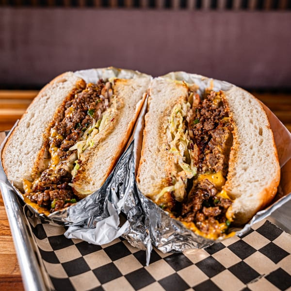A Clutch City Grill cheesesteak sandwich, cut in half, with melted cheese and meat, on a checkered paper.
