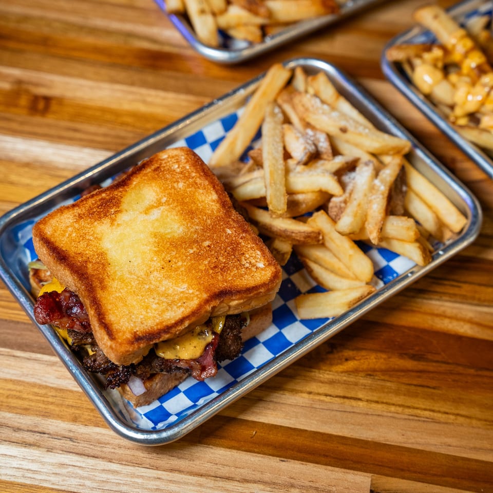 Clutch City Grill's patty melt with bacon, cheese, and grilled onions, served with fries on a metal tray.