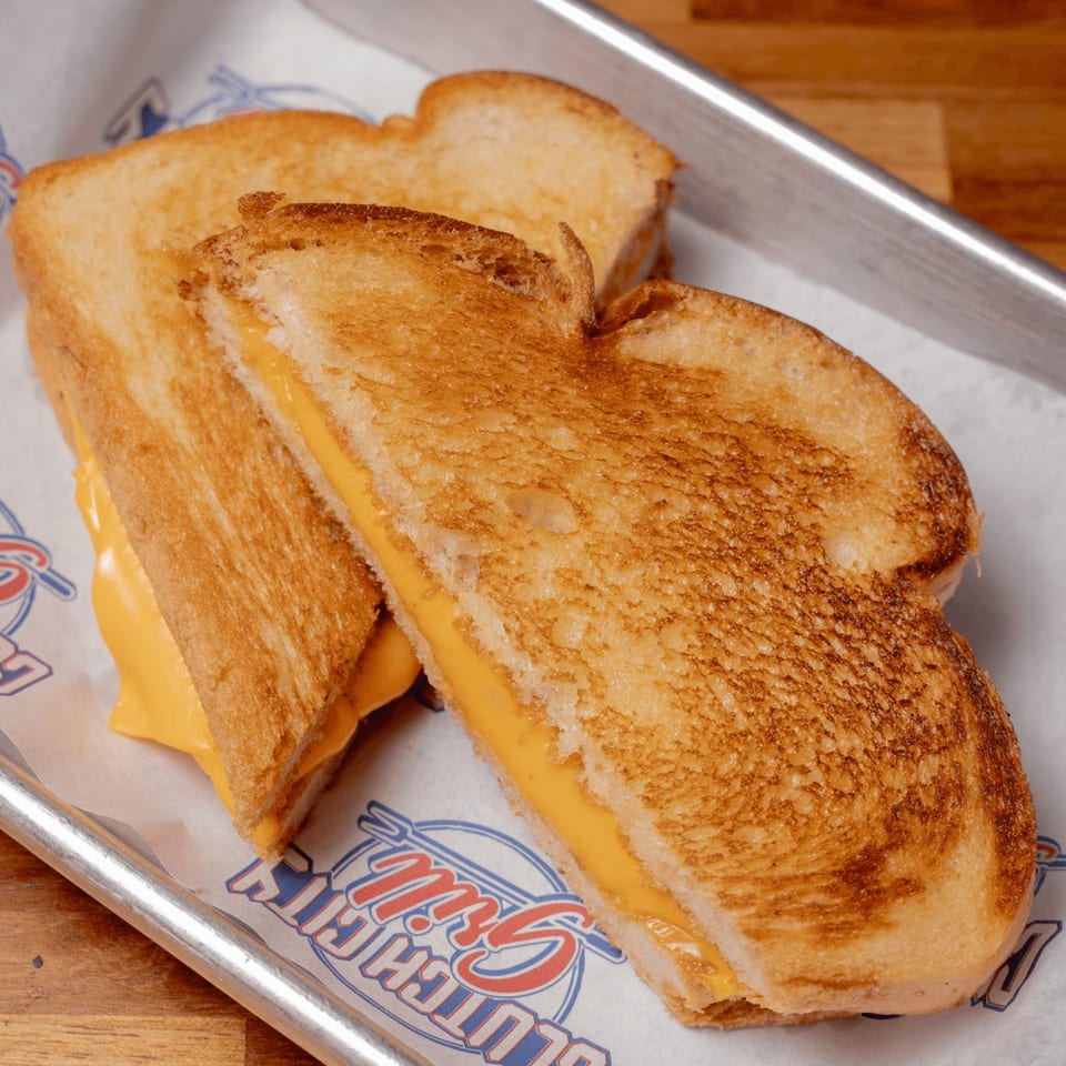 Two grilled cheese sandwiches with melted yellow cheese on a branded paper liner in a metal tray.
